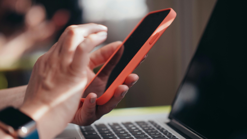Close-up of hands using a smartphone and laptop indoors, showcasing technology interaction.